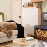 A cozy living room scene featuring a cream-colored sofa adorned with a fluffy beige fur pillow, a chunky knit beige throw blanket, and a dark brown faux fur throw. In front of the sofa is a round light beige stone coffee table holding a book and a small bowl.