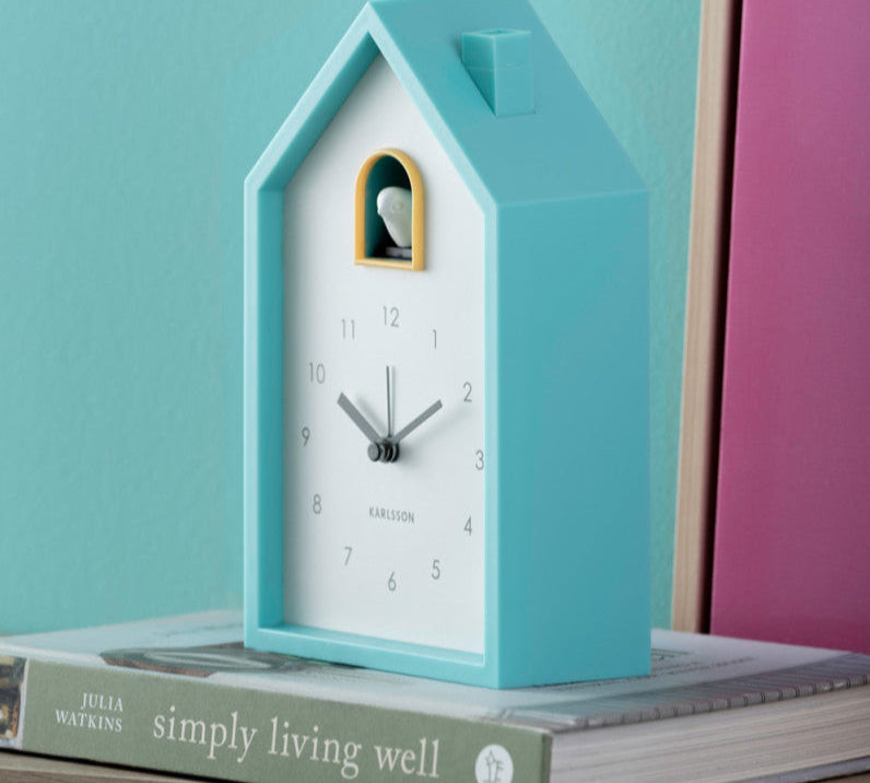 Blue cuckoo clock on a stack of books with a teal and pink background
