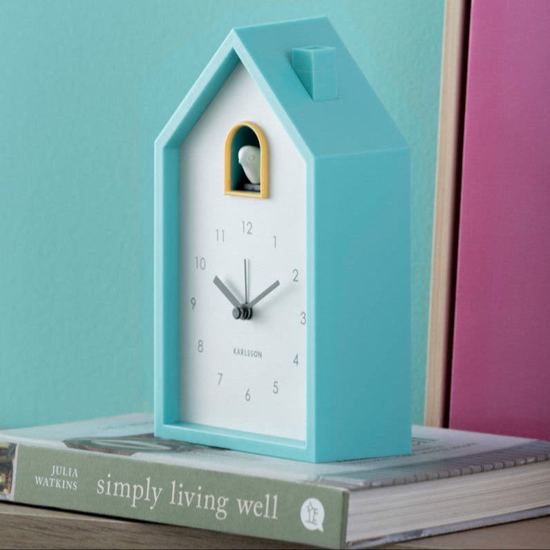 Blue cuckoo clock on a stack of books with a teal and pink background