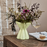 Green vase with flowers on a wooden table with a glass of wine and a muffin.