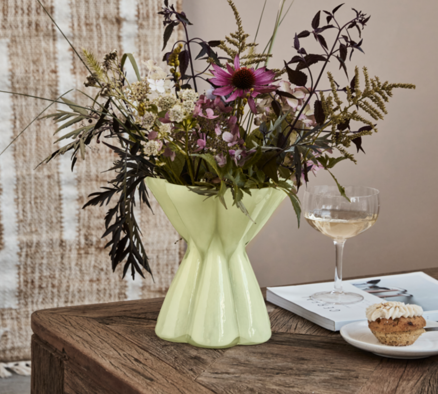 Green vase with flowers on a wooden table with a glass of wine and a muffin.