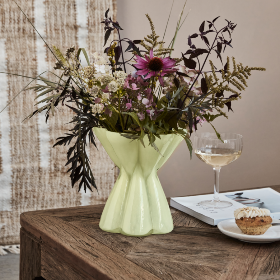 Green vase with flowers on a wooden table with a glass of wine and a muffin.