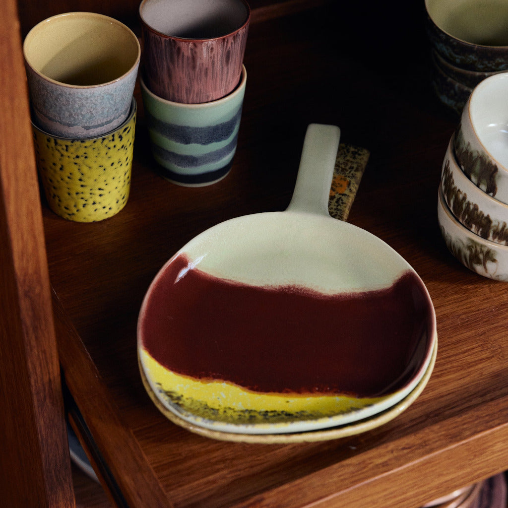 A ceramic serving bowl with a dark red and yellow marbled glaze sits on a wooden shelf, surrounded by other colorful stoneware cups and dishes.