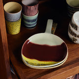 A ceramic serving bowl with a dark red and yellow marbled glaze sits on a wooden shelf, surrounded by other colorful stoneware cups and dishes.
