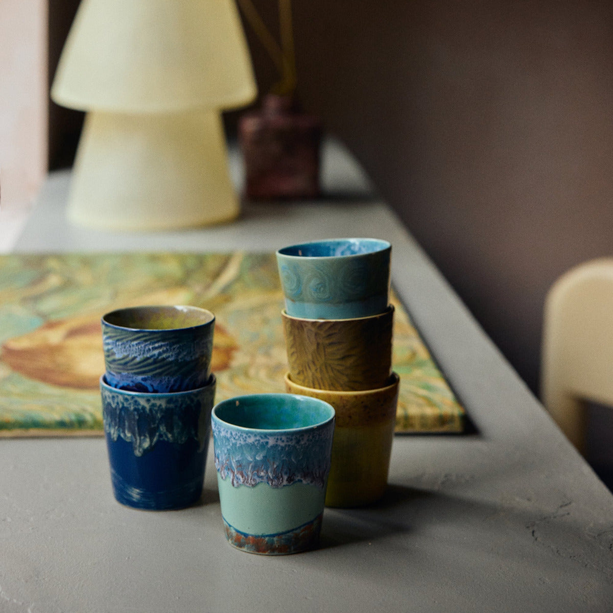 Stack of ceramic cups in blue and brown on a table with a lamp and painting in the background.