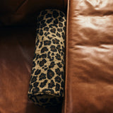 A cylindrical bolster pillow with a leopard print pattern, resting on a brown leather sofa.