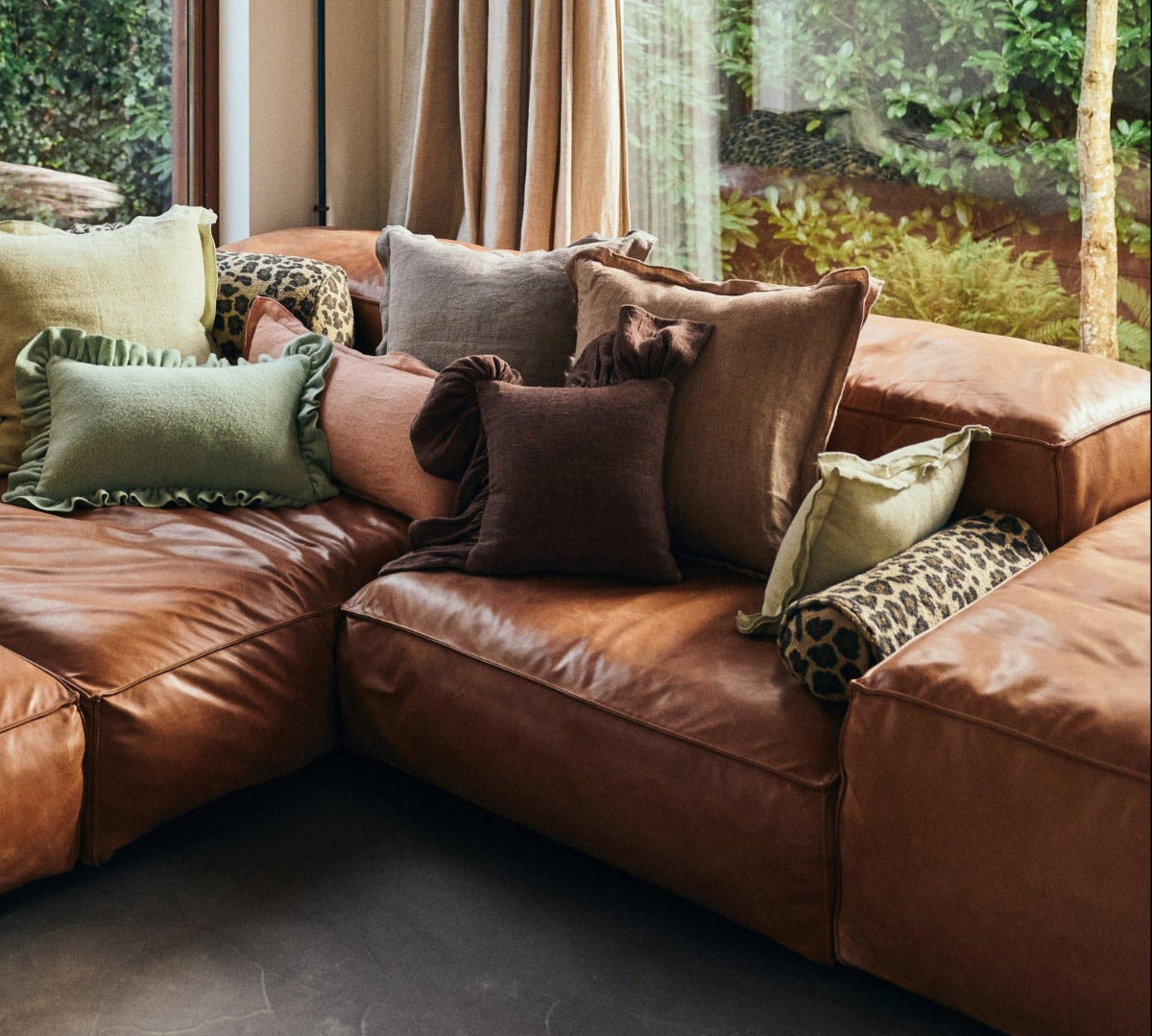 A large brown leather sectional sofa in a rustic living room with exposed wood ceiling beams, large windows showing a forest view, and multiple decorative throw pillows.