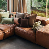 A large brown leather sectional sofa in a rustic living room with exposed wood ceiling beams, large windows showing a forest view, and multiple decorative throw pillows.