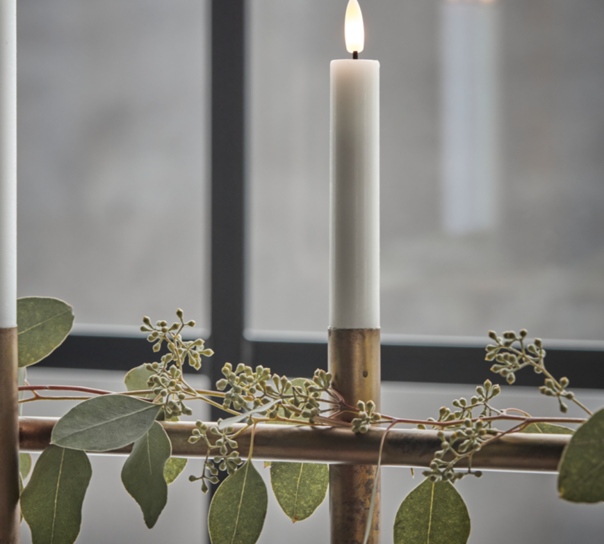 Two lit candles in wooden holders with greenery on a reflective surface.