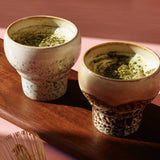 Two ceramic teacups with green tea on a wooden tray against a pink background