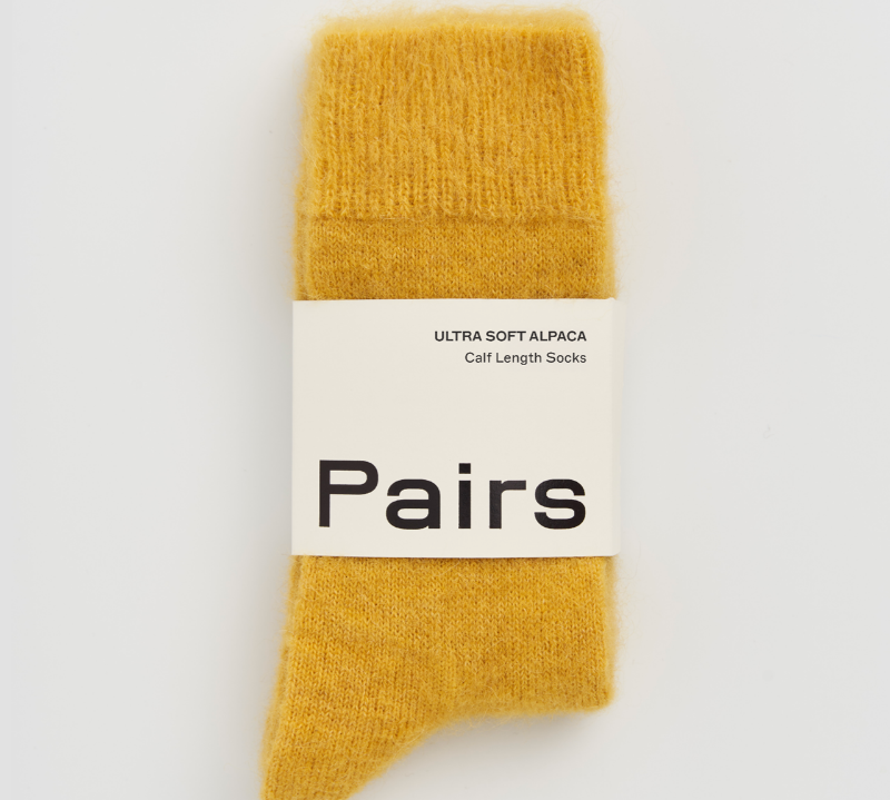 A pair of soft mustard yellow knit socks in their original packaging, featuring a white paper band with the brand name 'Pairs' printed in black.