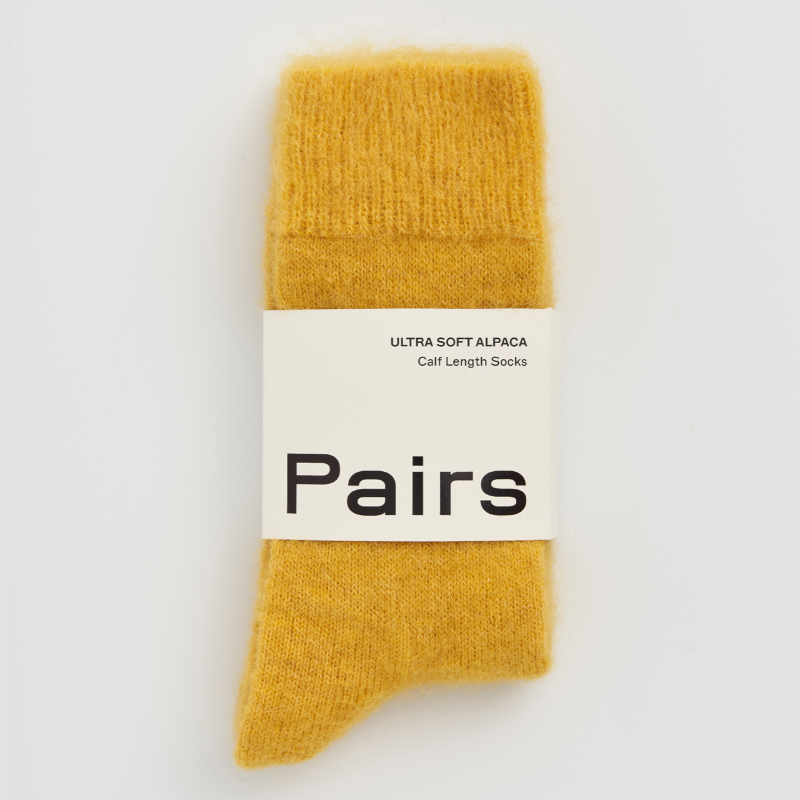 A pair of soft mustard yellow knit socks in their original packaging, featuring a white paper band with the brand name 'Pairs' printed in black.