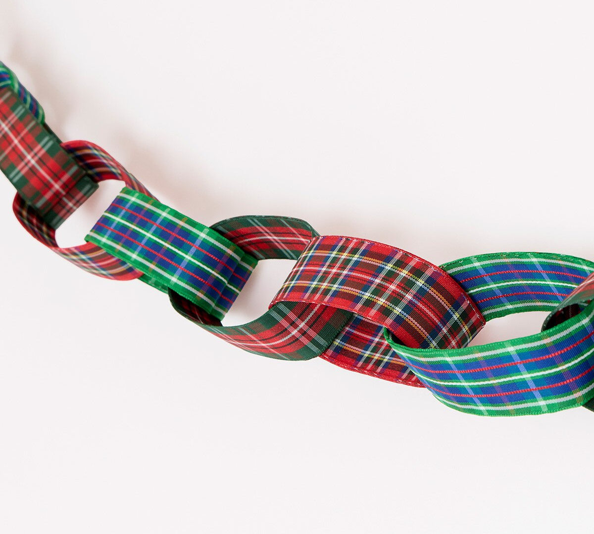 A close-up of a woven ribbon garland made of interlocking loops in a red, green, blue, and white tartan plaid pattern, photographed against a plain white background.