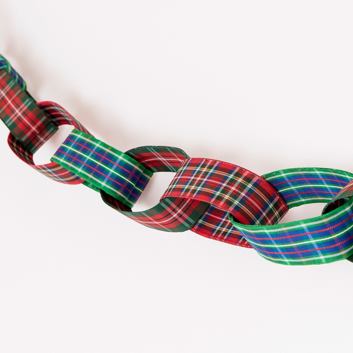 A close-up of a woven ribbon garland made of interlocking loops in a red, green, blue, and white tartan plaid pattern, photographed against a plain white background.