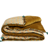 A folded quilted comforter with a rich mustard-gold velvet side and a patterned side featuring teal and orange botanical motifs, set against a plain white background.