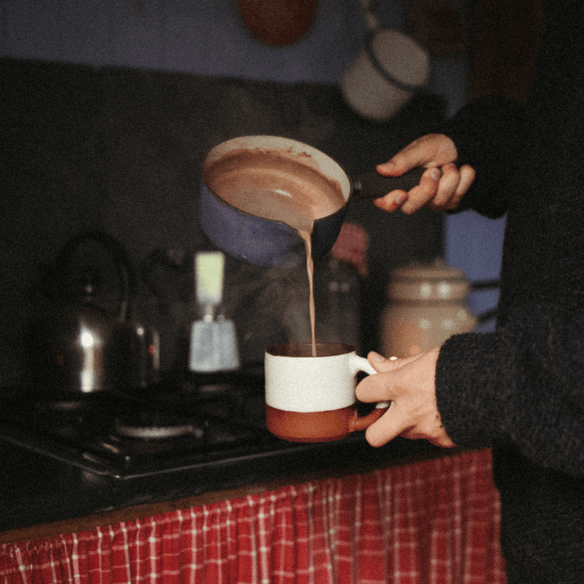 Person pouring hot chocolate from a pot into a mug on a stove.