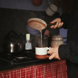 Person pouring hot chocolate from a pot into a mug on a stove.