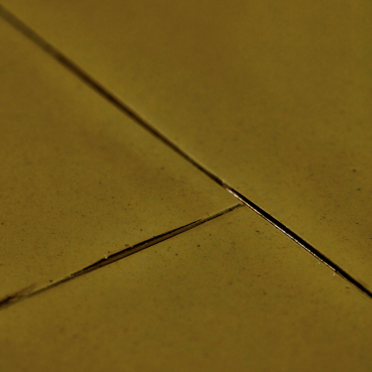 Close-up of a textured surface in warm olive-gold tones with thin dark grout or seam lines crossing diagonally.