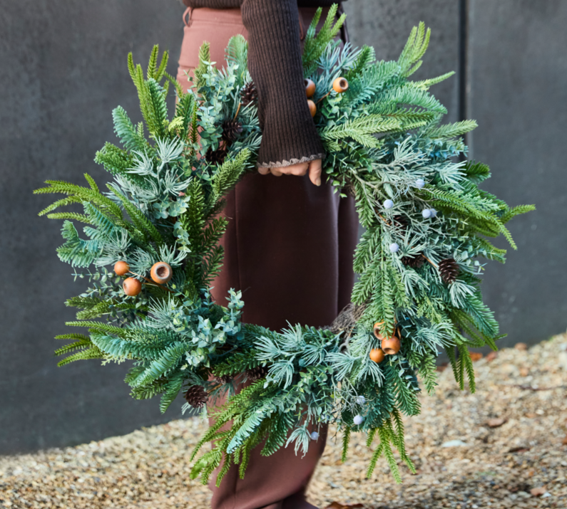 A person outdoors holding a large green Christmas wreath decorated with frosted pine branches, evergreen foliage, pine cones, and acorns. The person is wearing brown pants, a dark brown sweater, and tan boots, standing on gravel against a dark gray wall.