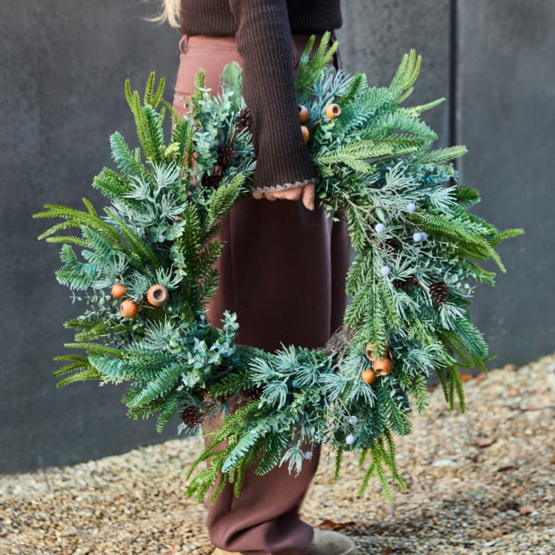 A person outdoors holding a large green Christmas wreath decorated with frosted pine branches, evergreen foliage, pine cones, and acorns. The person is wearing brown pants, a dark brown sweater, and tan boots, standing on gravel against a dark gray wall.