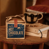 Box of hot chocolate on a wooden table with books and a mug in the background