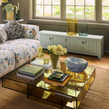 A modern square glass coffee table with a reflective amber-tinted glass top and black metal frame, styled in a bright living room with a floral sofa, books, and flowers.