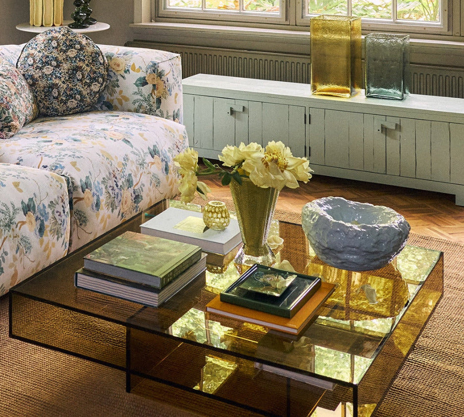 A modern square glass coffee table with a reflective amber-tinted glass top and black metal frame, styled in a bright living room with a floral sofa, books, and flowers.