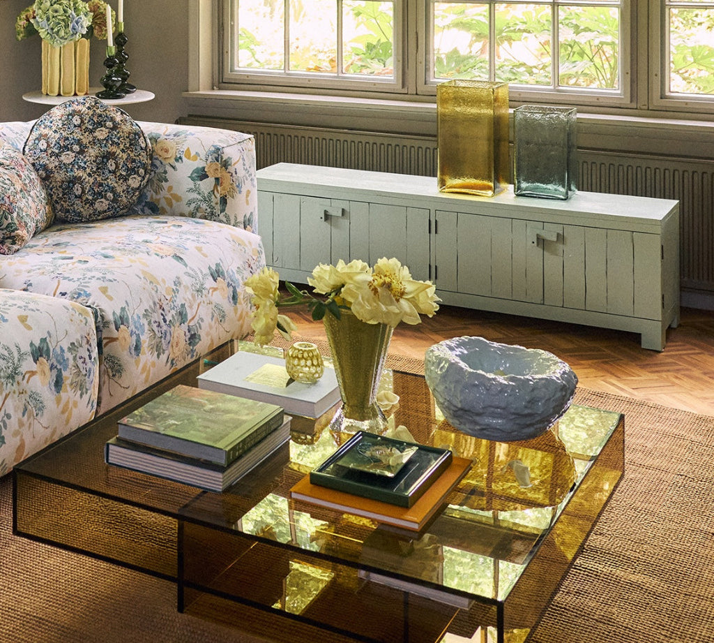 A modern square glass coffee table with a reflective amber-tinted glass top and black metal frame, styled in a bright living room with a floral sofa, books, and flowers.