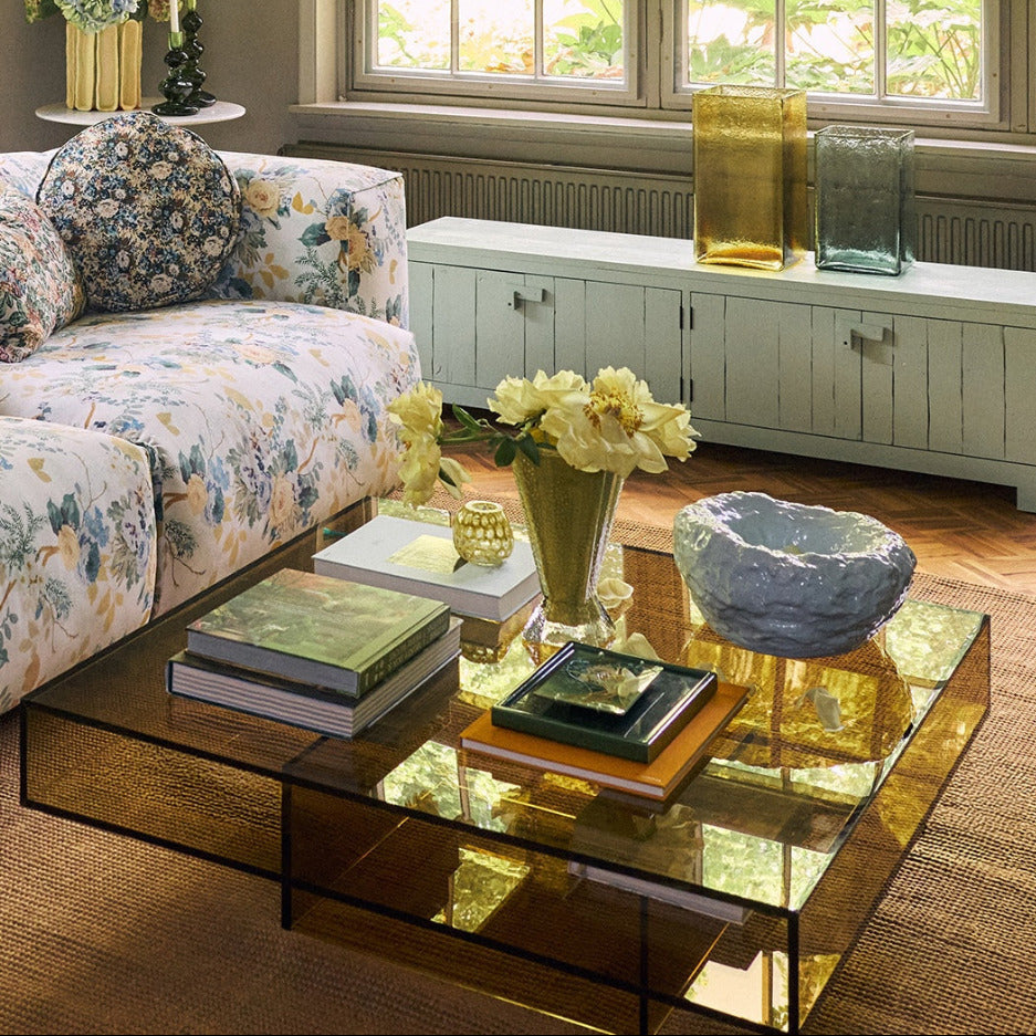 A modern square glass coffee table with a reflective amber-tinted glass top and black metal frame, styled in a bright living room with a floral sofa, books, and flowers.