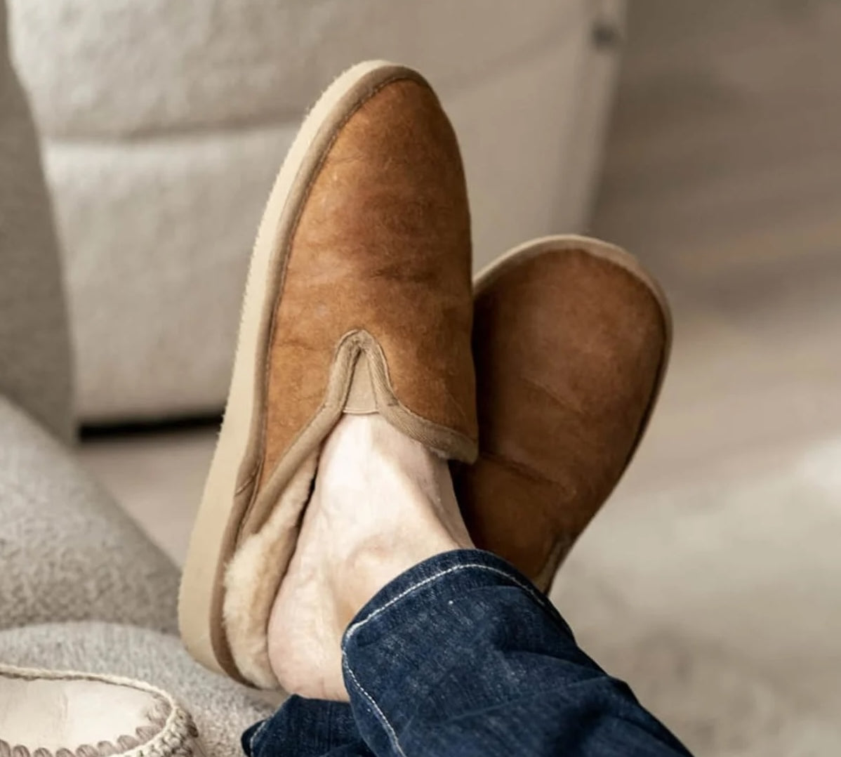A person wearing a pair of brown suede slip-on slippers with a cream sherpa lining, shown with blue jeans in a cozy indoor setting.