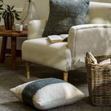 A styled living room scene featuring a cream upholstered armchair with a dark gray textured throw pillow, a matching patterned lumbar pillow on the floor, a wooden side table with a glass lamp, and a wicker basket.