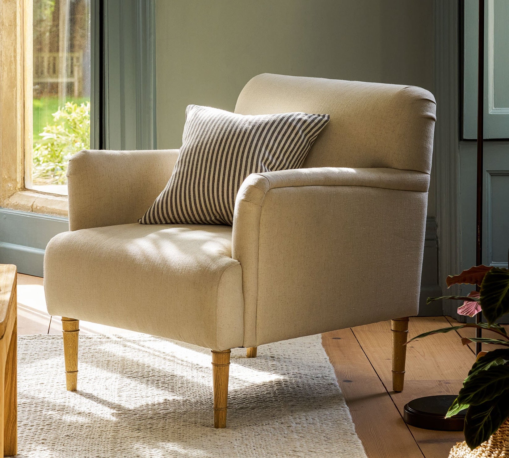 A cream-colored upholstered armchair with a striped throw pillow, set in a sunlit room with a window, wooden floor, and a black floor lamp.
