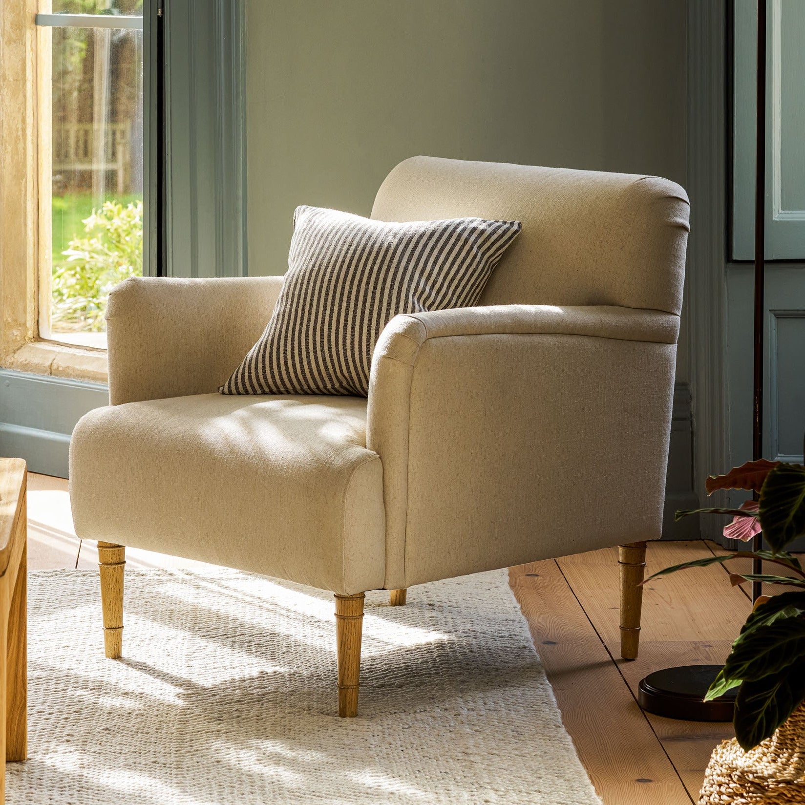 A cream-colored upholstered armchair with a striped throw pillow, set in a sunlit room with a window, wooden floor, and a black floor lamp.