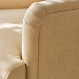 Close-up of a beige upholstered armchair showing the fabric texture and rounded arm detail in warm natural light.