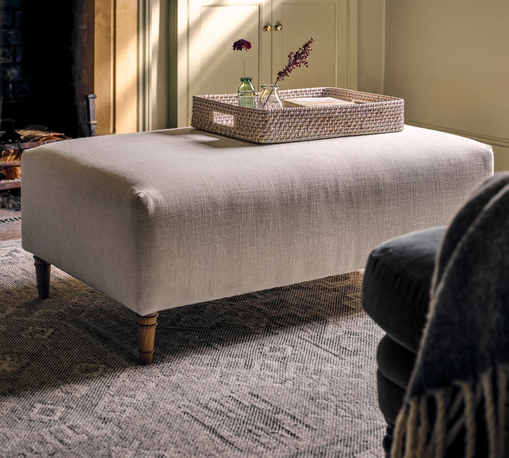 A light beige upholstered ottoman with turned wooden legs stands in a styled living room in front of a fireplace and built-in shelves, with a woven tray resting on top.