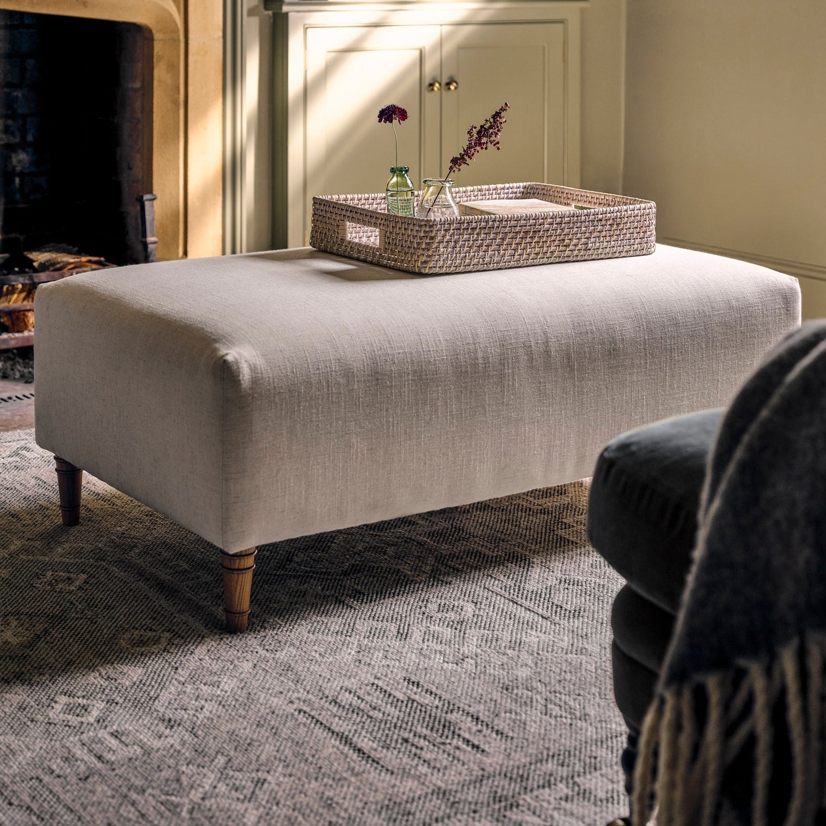 A light beige upholstered ottoman with turned wooden legs stands in a styled living room in front of a fireplace and built-in shelves, with a woven tray resting on top.