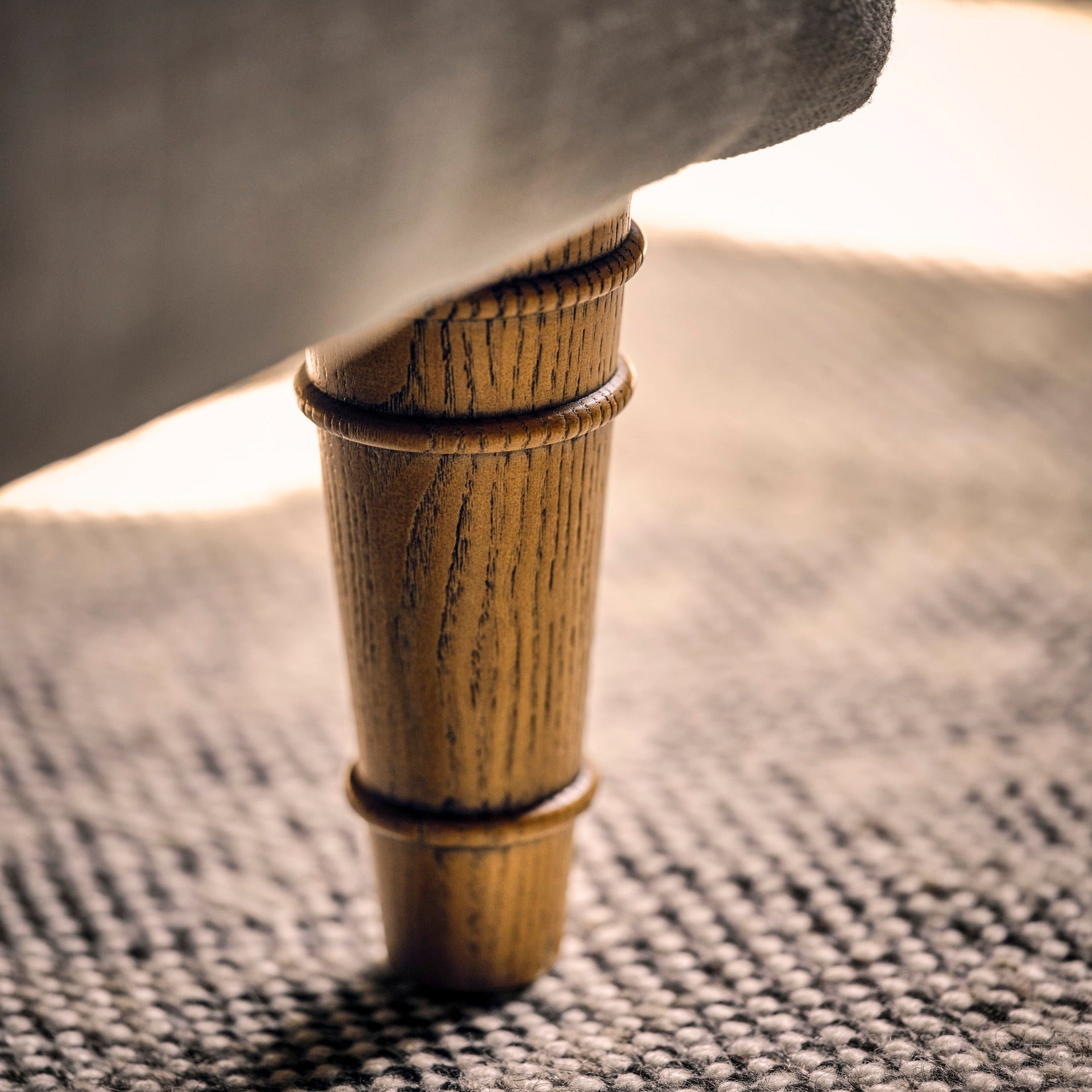 Close-up of a wooden upholstered chair leg resting on a textured gray rug, with warm sunlight and soft shadows.
