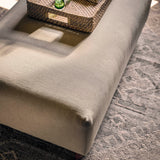 A large upholstered ottoman or bench in a light warm gray-beige fabric, styled with a woven tray, books, and small green glass vases on top, set on a patterned rug in a sunlit room.
