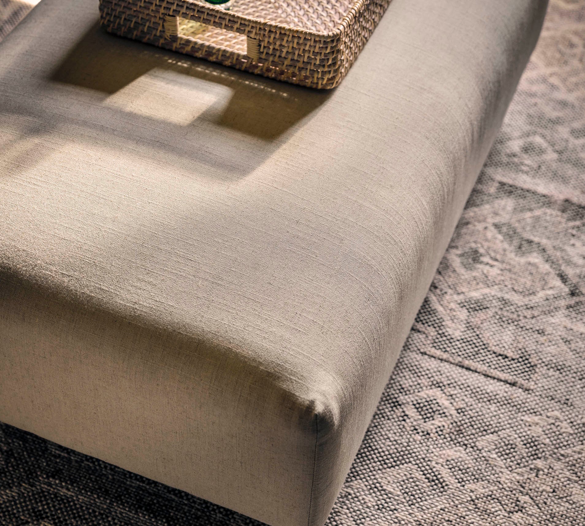 A large upholstered ottoman or bench in a light warm gray-beige fabric, styled with a woven tray, books, and small green glass vases on top, set on a patterned rug in a sunlit room.
