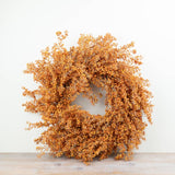A circular autumnal floral wreath in warm orange-brown tones, displayed against a plain off-white wall on a light wood surface.