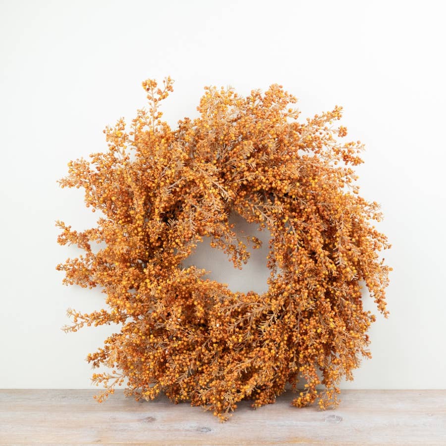 A circular autumnal floral wreath in warm orange-brown tones, displayed against a plain off-white wall on a light wood surface.