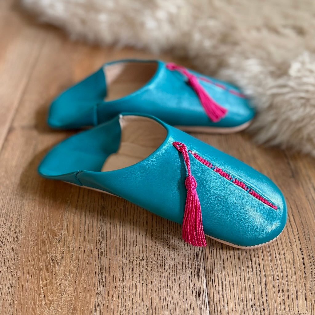 Pair of blue slippers with pink tassels on a wooden floor.