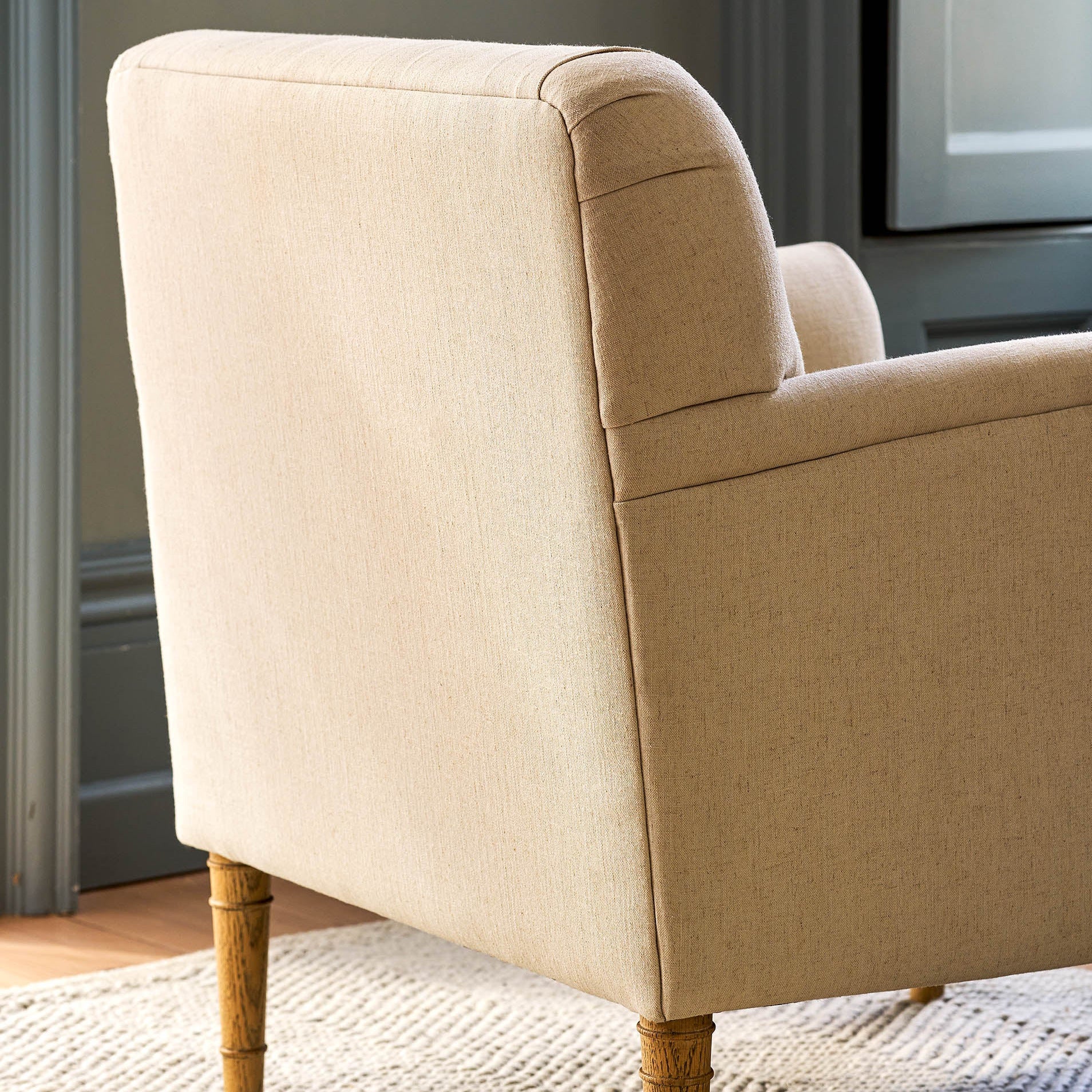 Close-up of the back and side of a beige upholstered armchair with turned wood legs, placed on a woven rug in an indoor room setting.