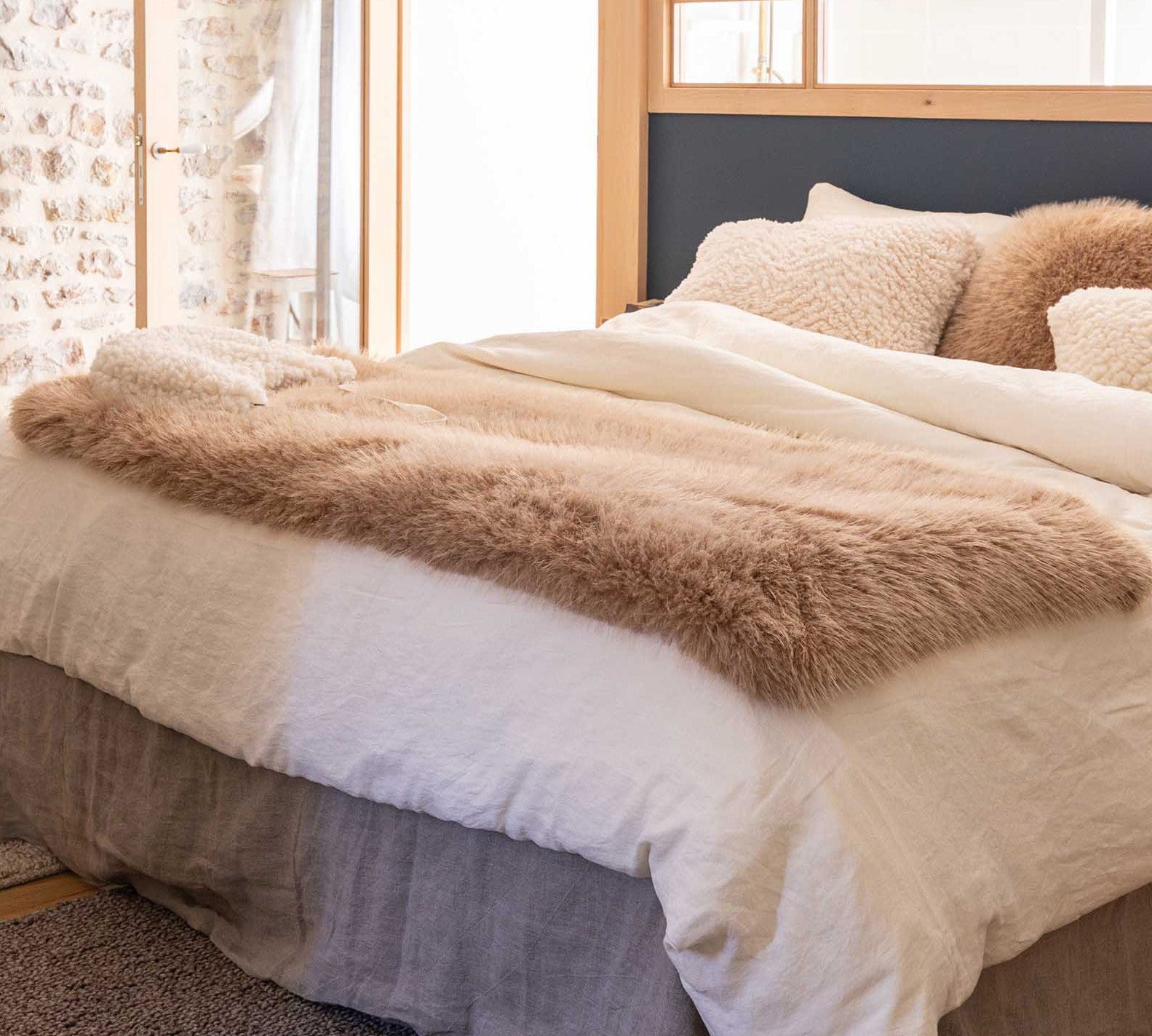 A neatly made bed in a bright bedroom, styled with cream bedding, textured beige pillows, a fluffy tan throw blanket, and a dark navy headboard.
