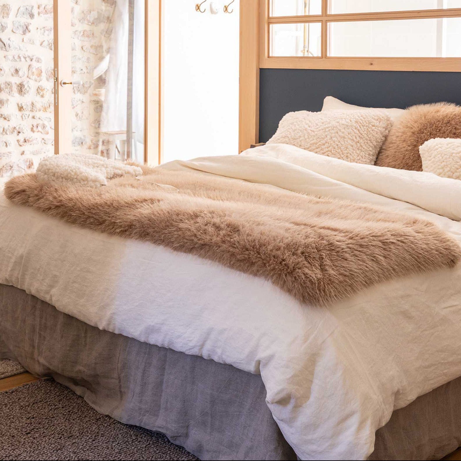 A neatly made bed in a bright bedroom, styled with cream bedding, textured beige pillows, a fluffy tan throw blanket, and a dark navy headboard.