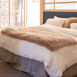 A neatly made bed in a bright bedroom, styled with cream bedding, textured beige pillows, a fluffy tan throw blanket, and a dark navy headboard.
