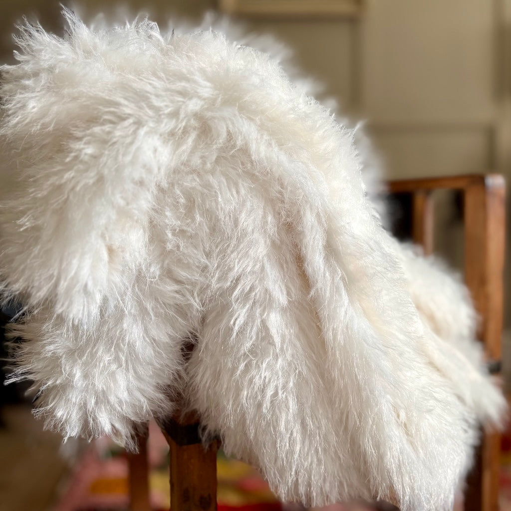 A fluffy white sheepskin throw blanket draped over a wooden chair in a softly blurred indoor setting.