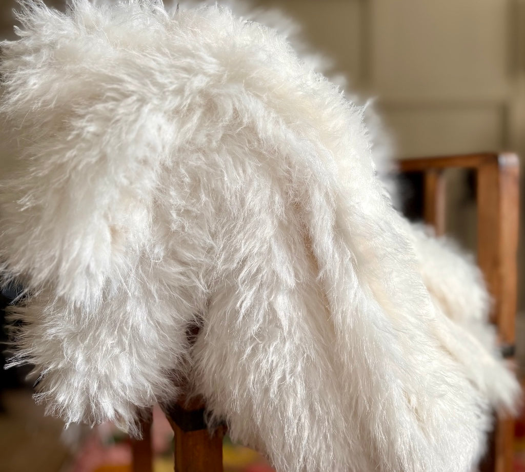 A fluffy white sheepskin throw blanket draped over a wooden chair in a softly blurred indoor setting.