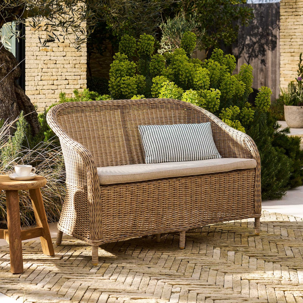 Rattan outdoor sofa with cushions on a patio area with plants and a brick wall in the background.