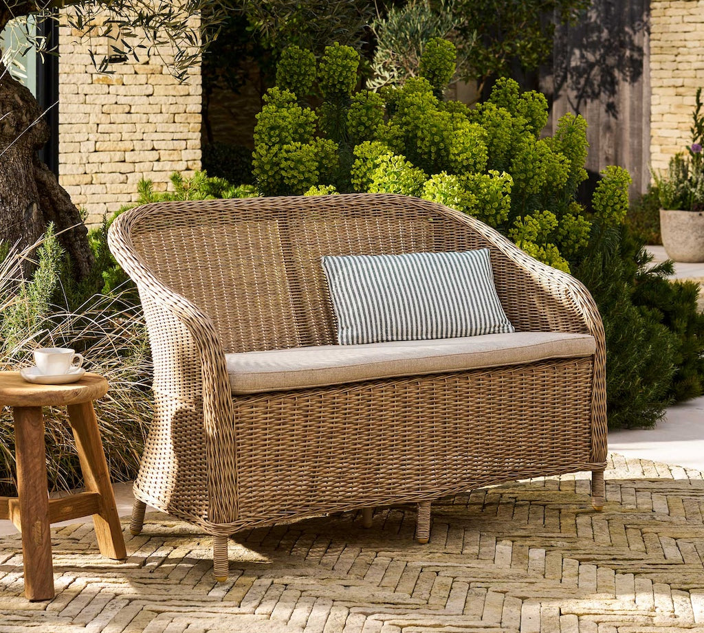 Rattan outdoor sofa with cushions on a patio area with plants and a brick wall in the background.